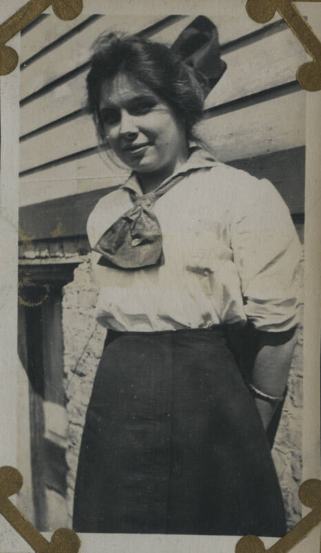 Untitled [Woman stands in front of house, arms behind back]