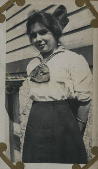 Untitled [Woman stands in front of house, arms behind back]