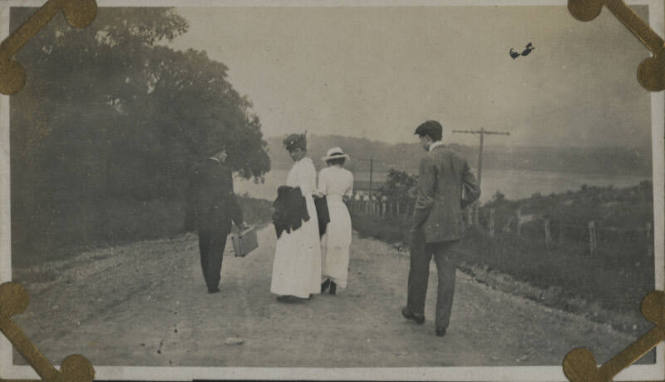 Untitled [Four people walk down dirt road, body of water in the distance]